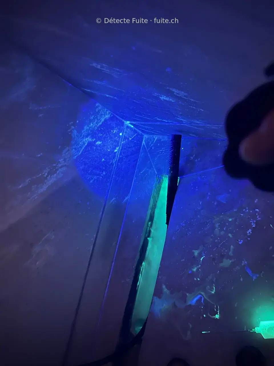 Surface intérieure éclairée sous UV, avec des traces fluorescentes près d’un passage de conduite